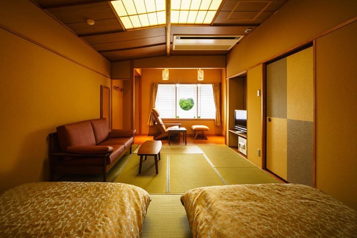 Ein traditioneller japanischer Raum mit Tatami-Bodenbelag, einer Futon-Liege, einem niedrigen Tisch, einer Couch und Schiebetüren.
