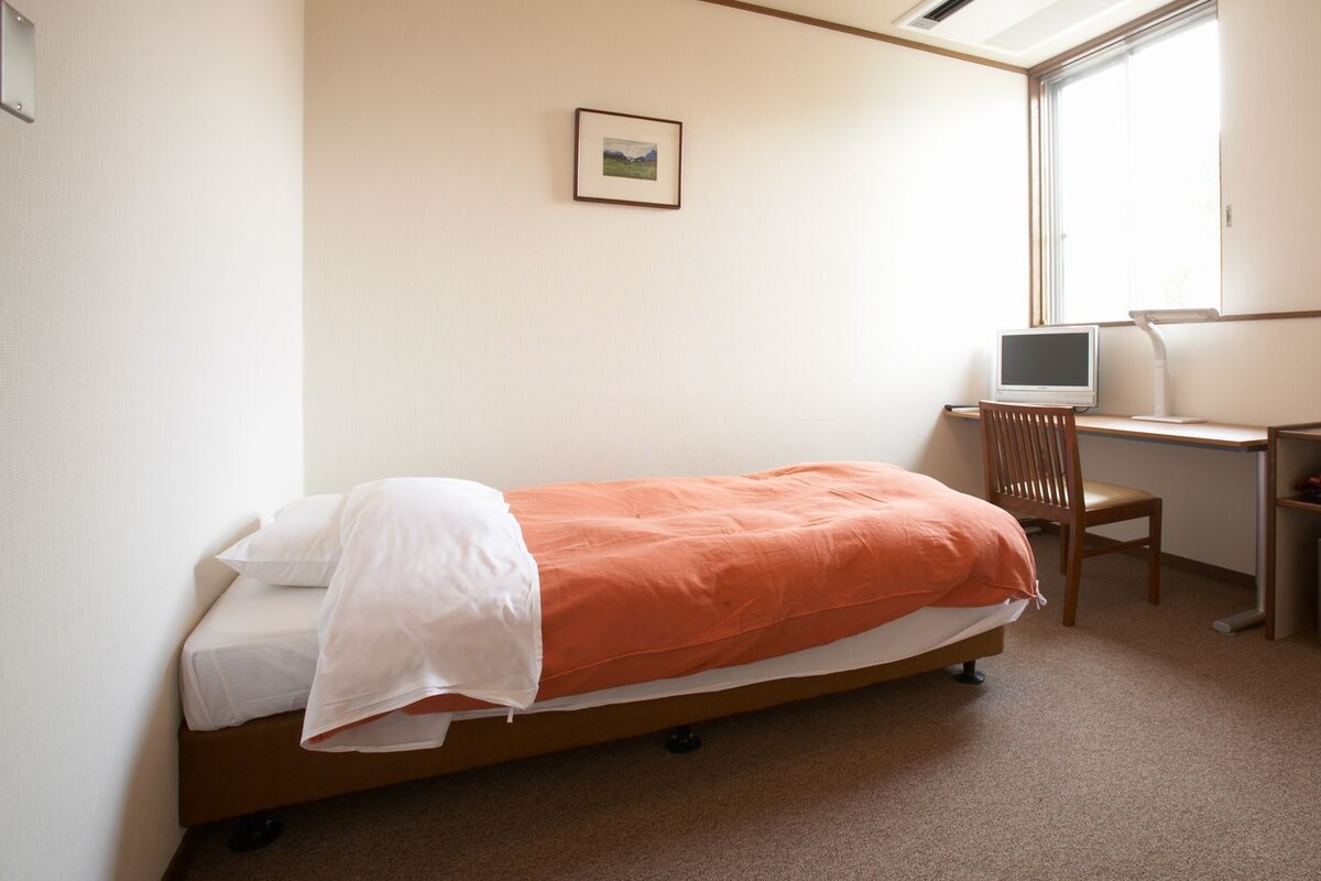 A single bed with an orange blanket, a desk with a computer, a chair, and a small cabinet.