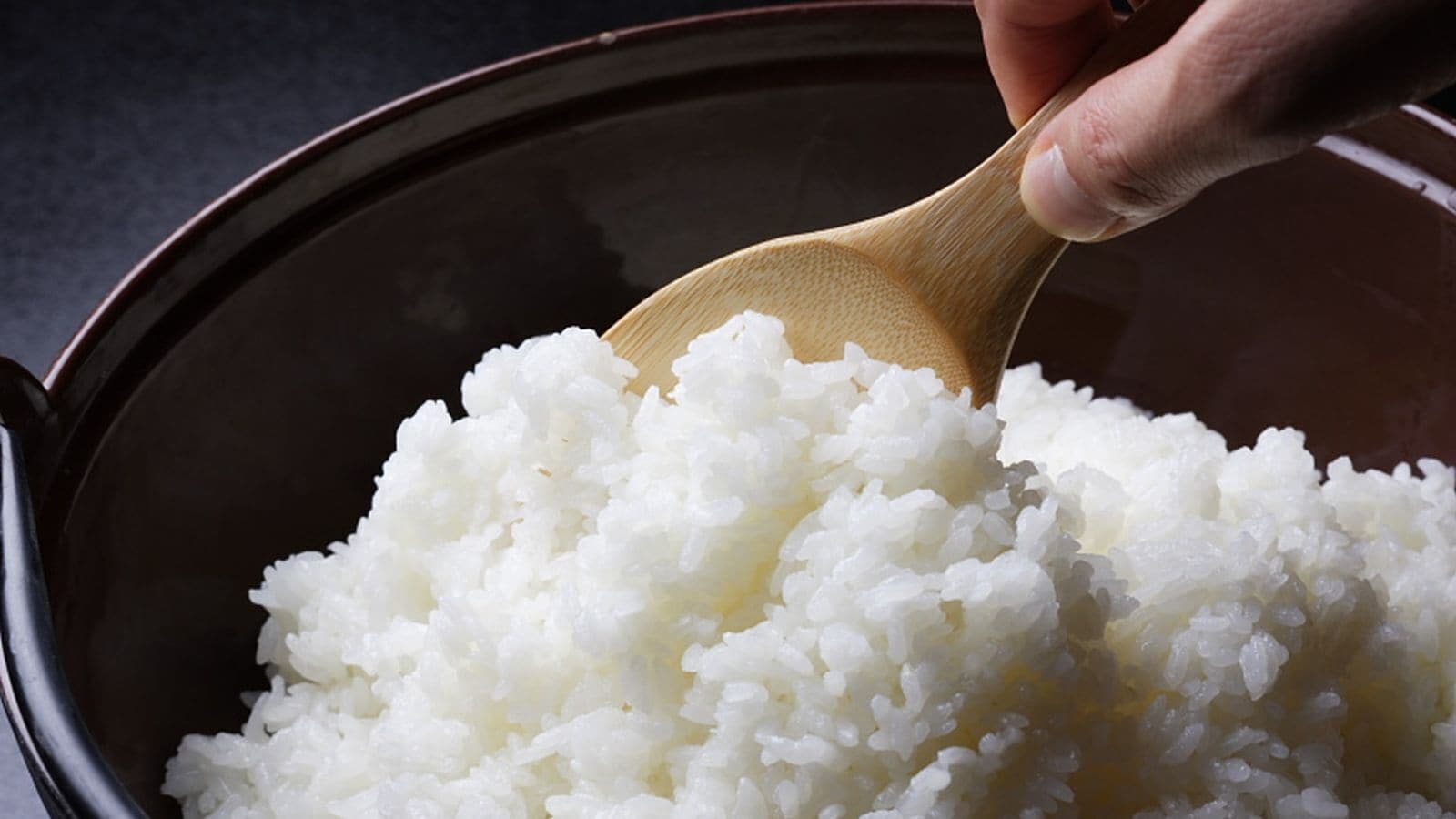 A hand holding a wooden spoon over a pot of cooked white rice.