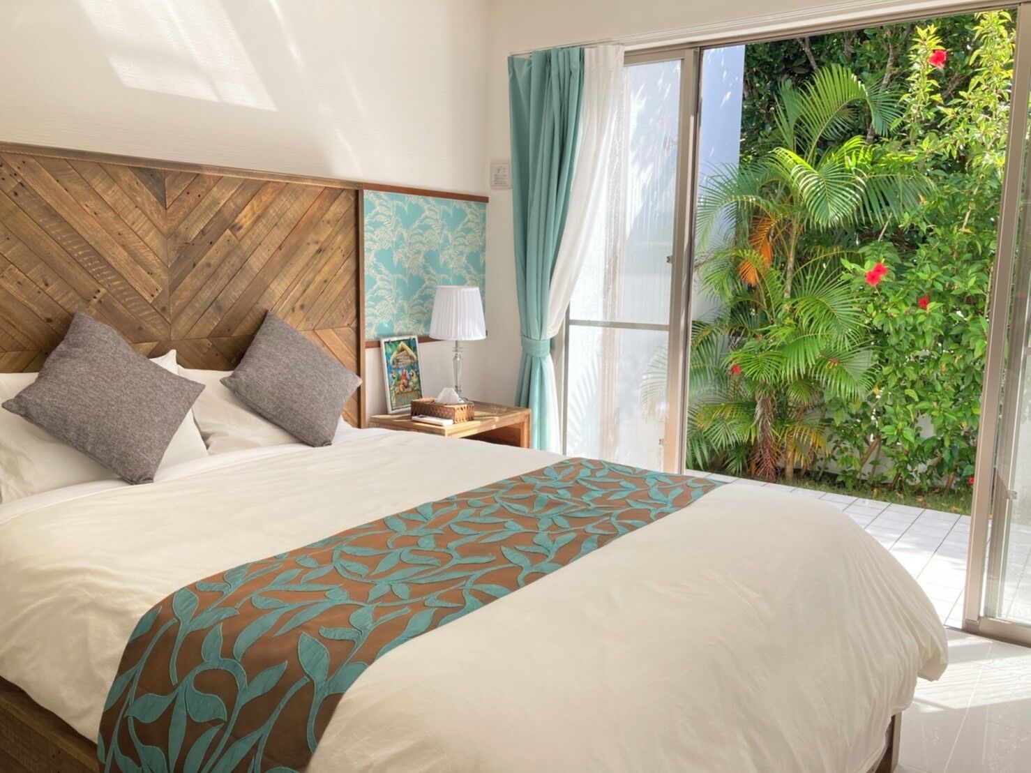 A hotel room with a large bed, a wooden headboard, a nightstand with a lamp, and a view of a lush garden through a sliding glass door.