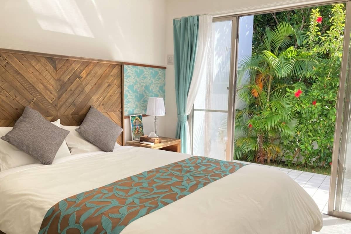 A hotel room with a large bed, a wooden headboard, a nightstand with a lamp, and a view of a lush garden through a sliding glass door.