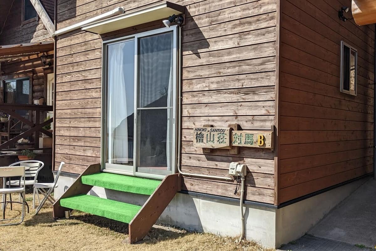 Una construcción de madera con puerta corredera de vidrio, un pequeño espacio de estar al aire libre y un letrero con caracteres japoneses.