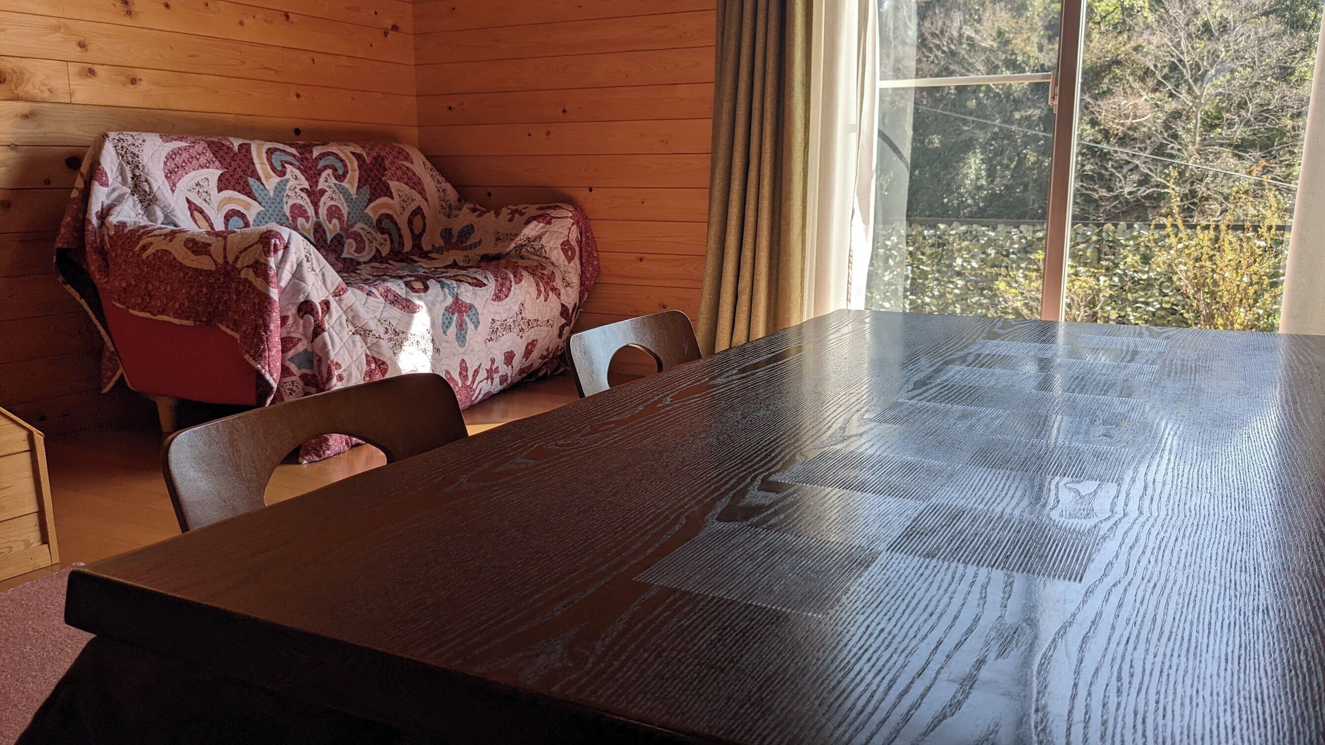 A wooden cabin interior with a large table, a patterned sofa, and a window with a view of trees.