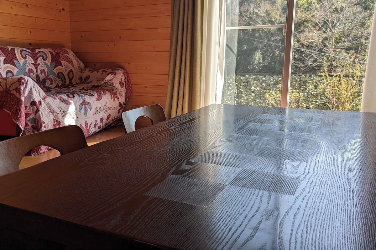 Interior de una cabaña de madera con una mesa grande, un sofá estampado y un ventanal con vistas a árboles.