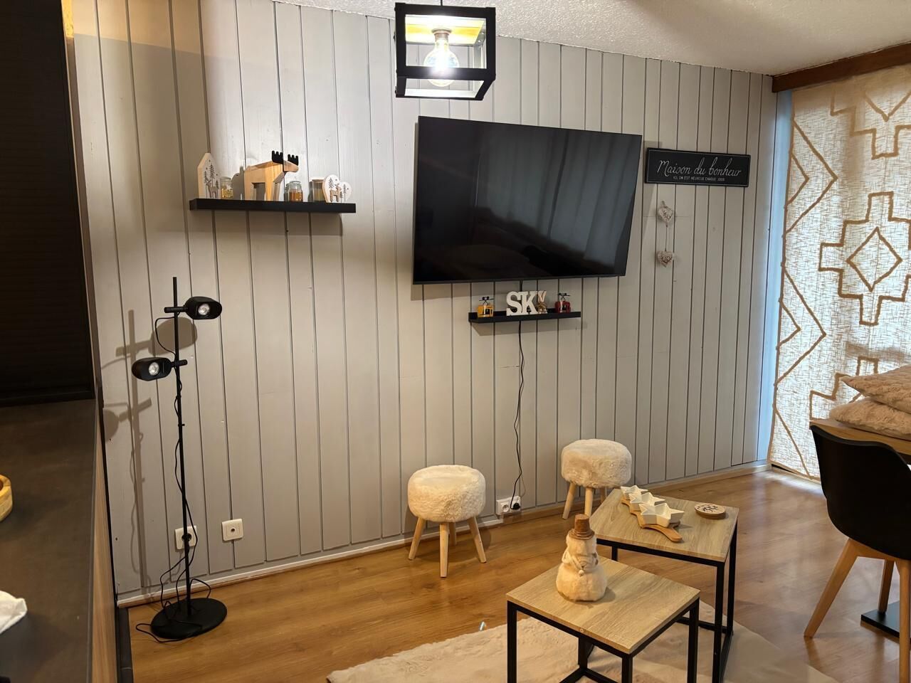 Un salon doté d’un téléviseur à écran plat, d’un parquet, de deux poufs, d’une petite table agrémentée d’objets décoratifs et d’une étagère fixée au mur.