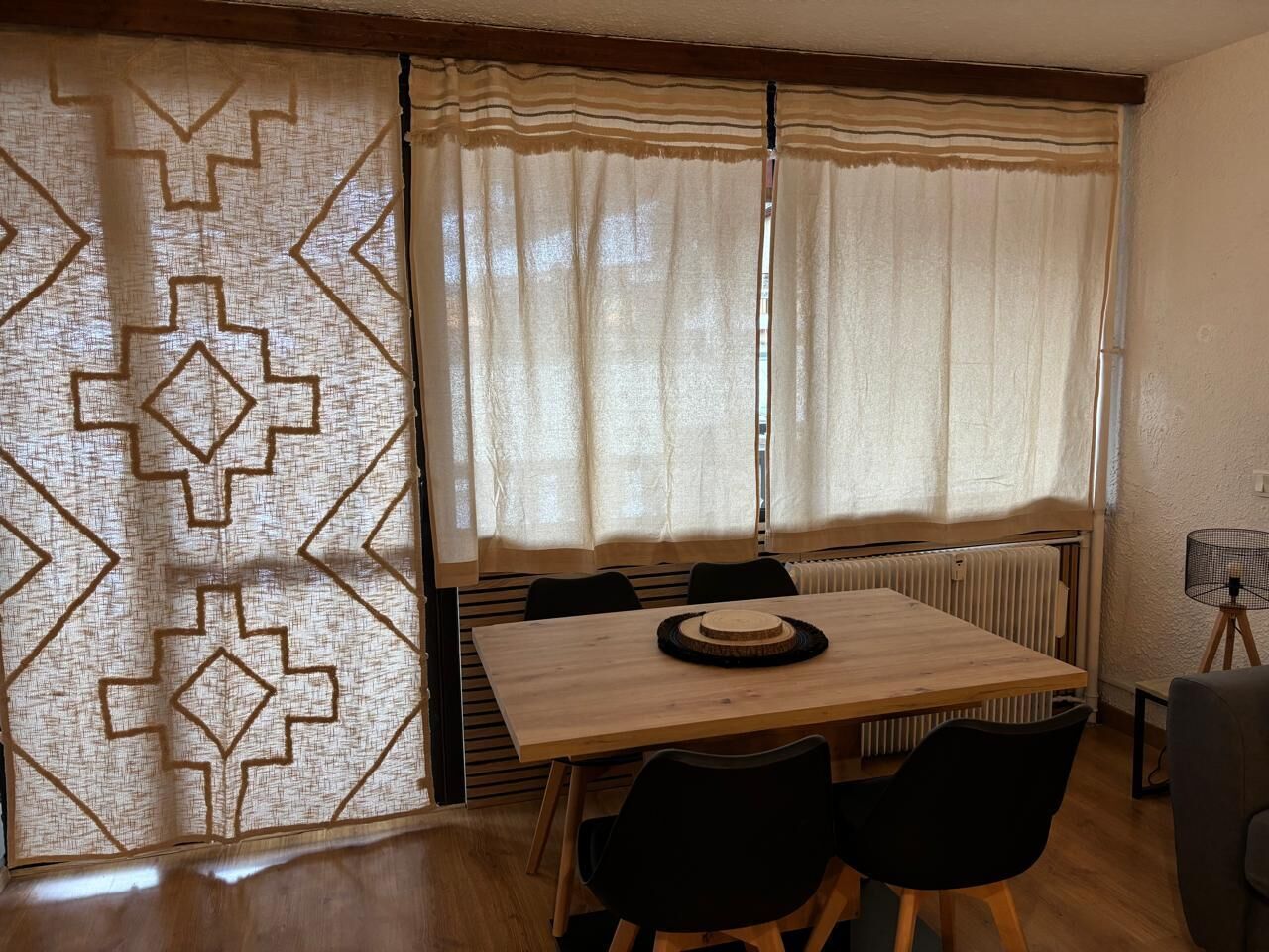 Un coin repas avec une table en bois, des chaises noires, un set de table en fibres tressées et un radiateur.