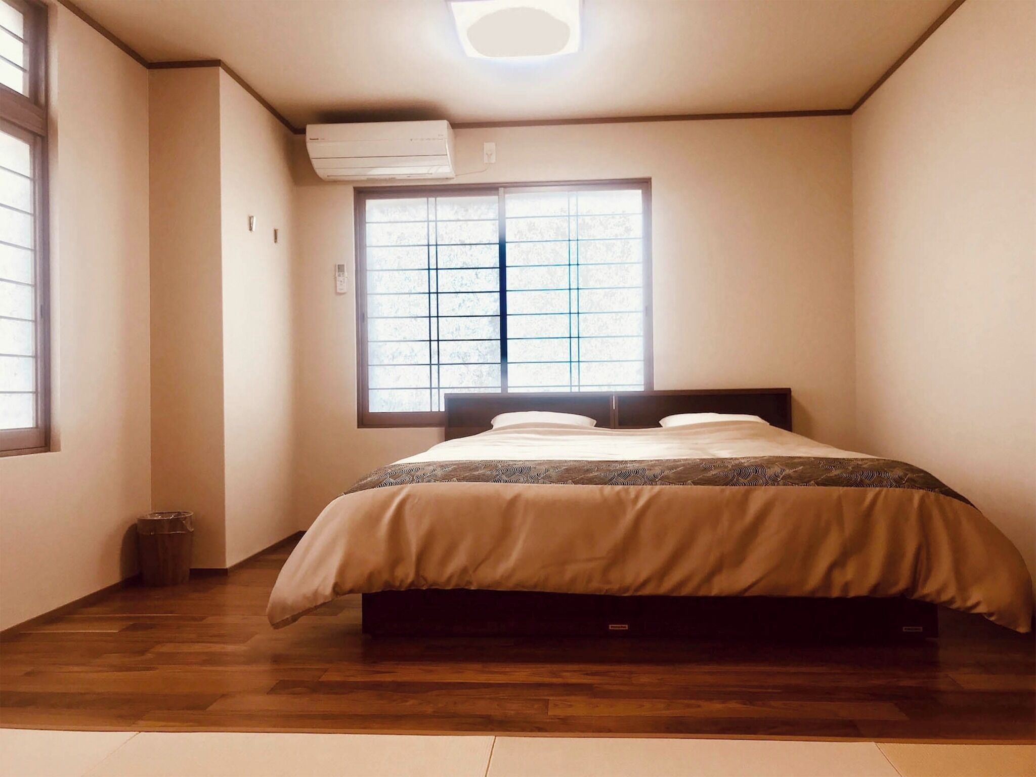 A bedroom with a bed, a window with blinds, an air conditioning unit, and a wooden floor.