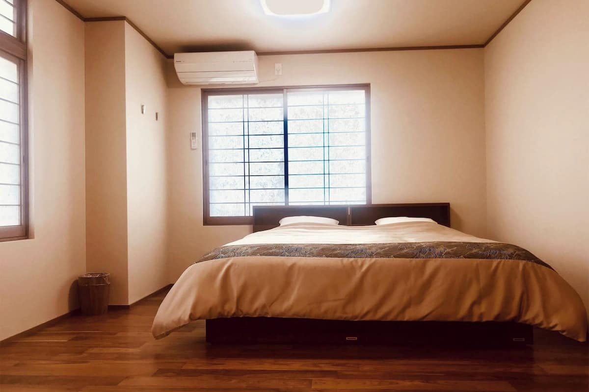 A bedroom with a bed, a window with blinds, an air conditioning unit, and a wooden floor.