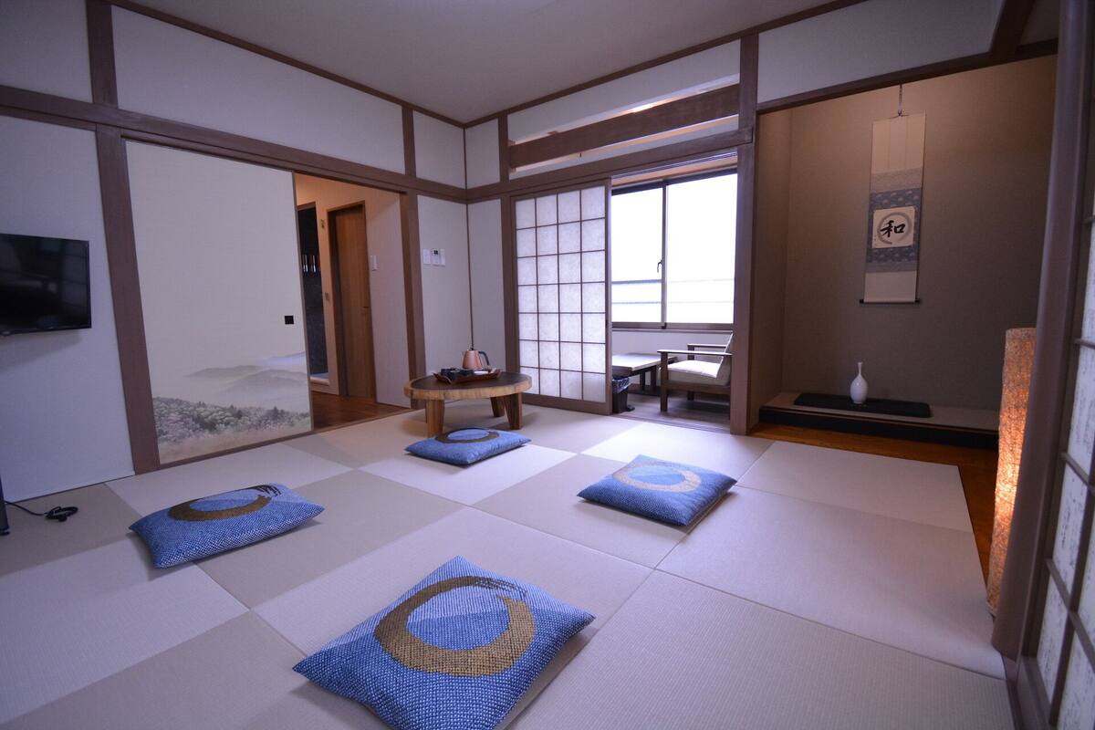 Een traditionele Japanse kamer met tatami vloerbedekking, een lage tafel met waterkoker, een stoel en een klein tafeltje met een vaas.