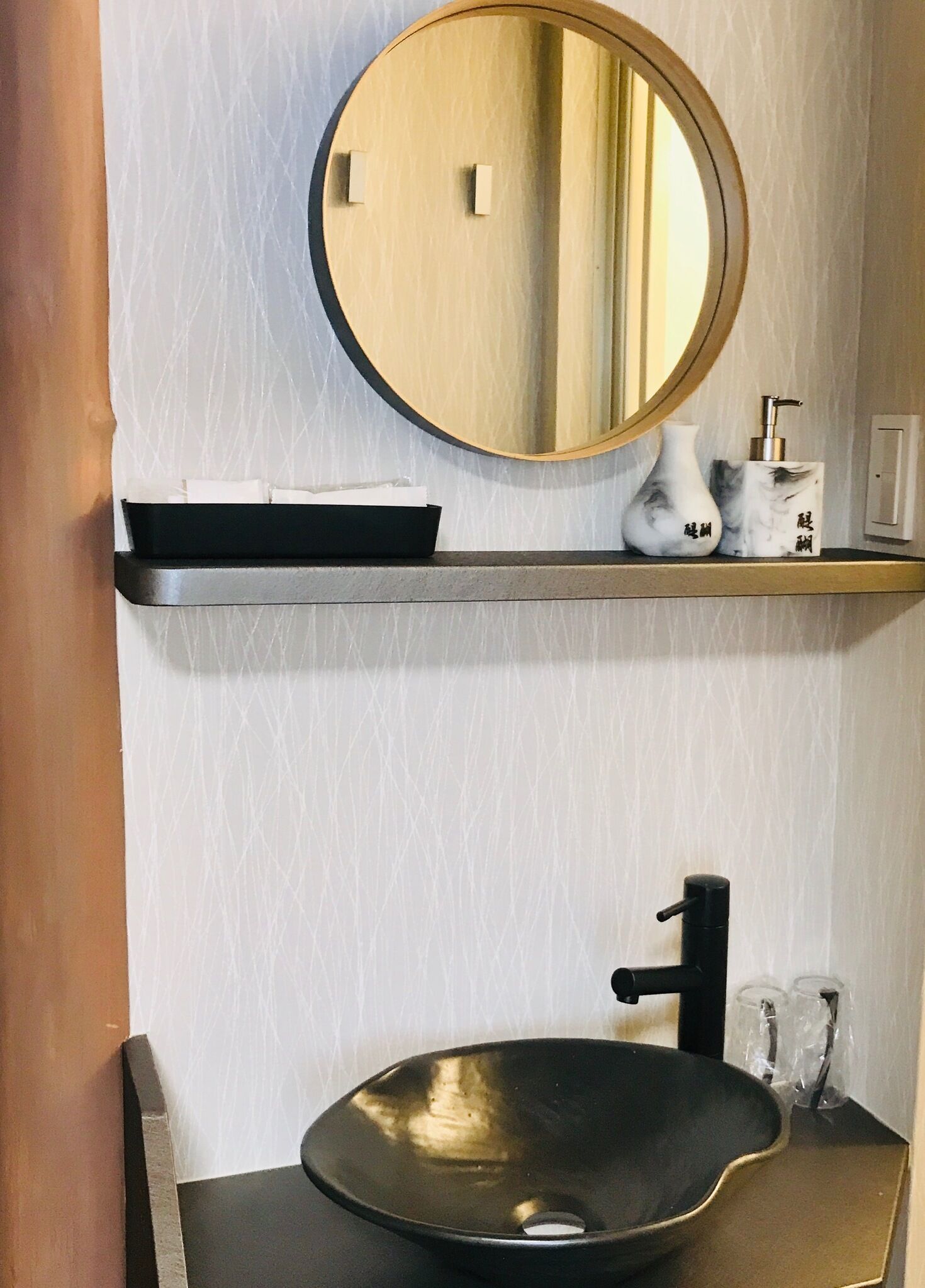 A bathroom with a round mirror, a black sink, and a shelf with a soap dispenser and a vase.