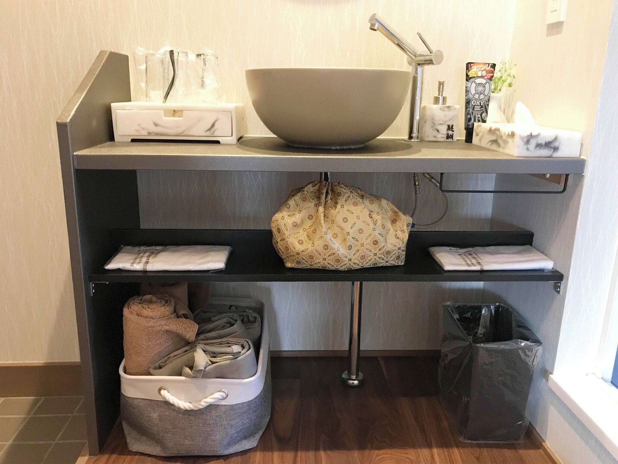 A bathroom with a large bowl sink, a towel rack, a trash bin, and a storage bin with towels.