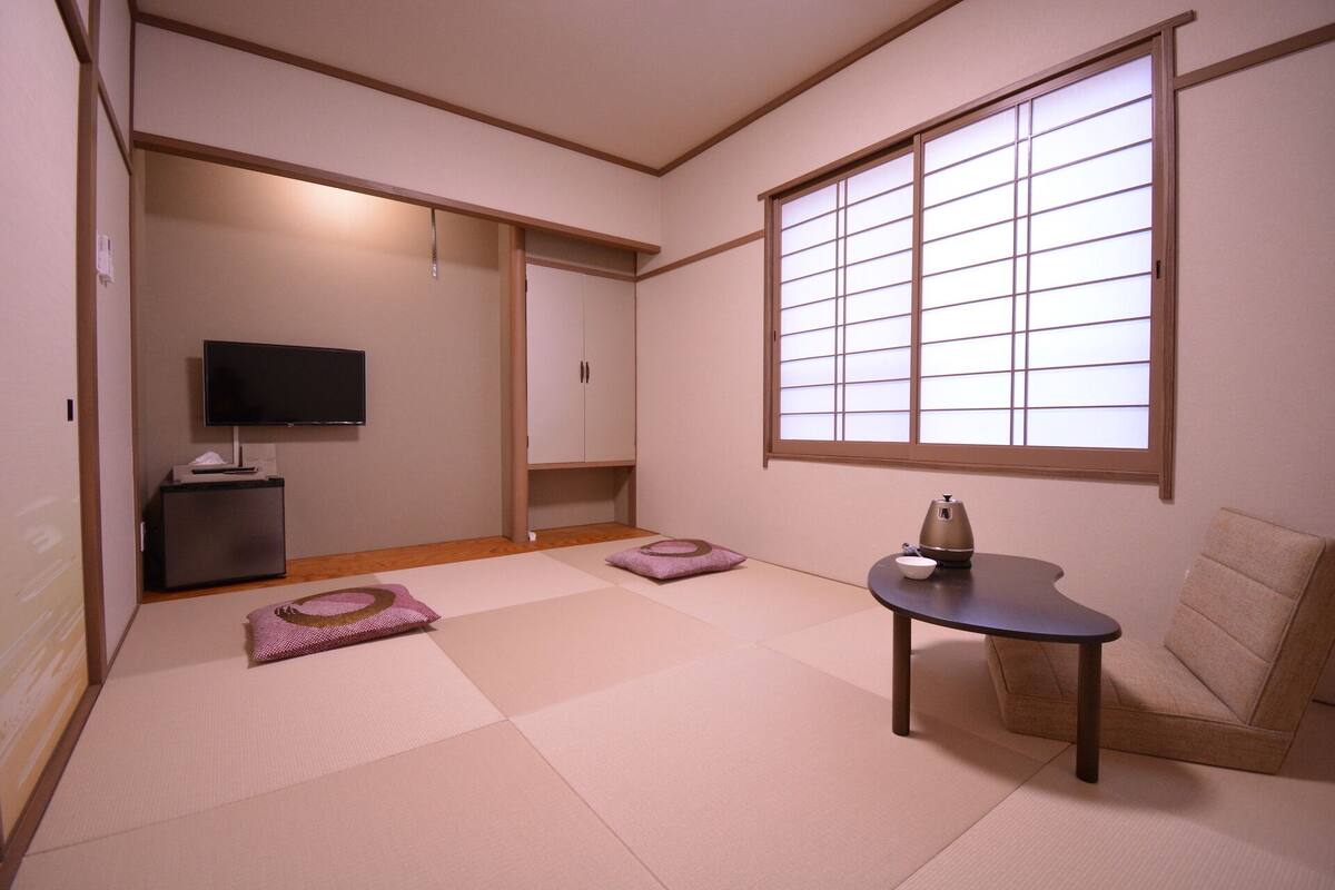 Een kamer met tatami vloer, een televisie, een kast, een tafel met waterkoker en een raam met shoji schermen.
