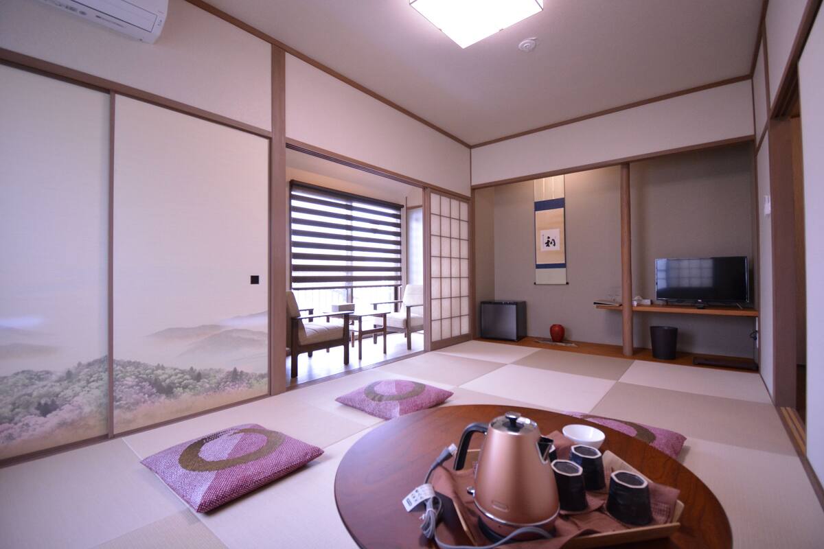 Een traditionele Japanse kamer met tatami vloeren, een tafel met tatami mat waarop een waterkoker en kopjes staan, een televisie en schuifdeuren die uitkomen op een balkon.
