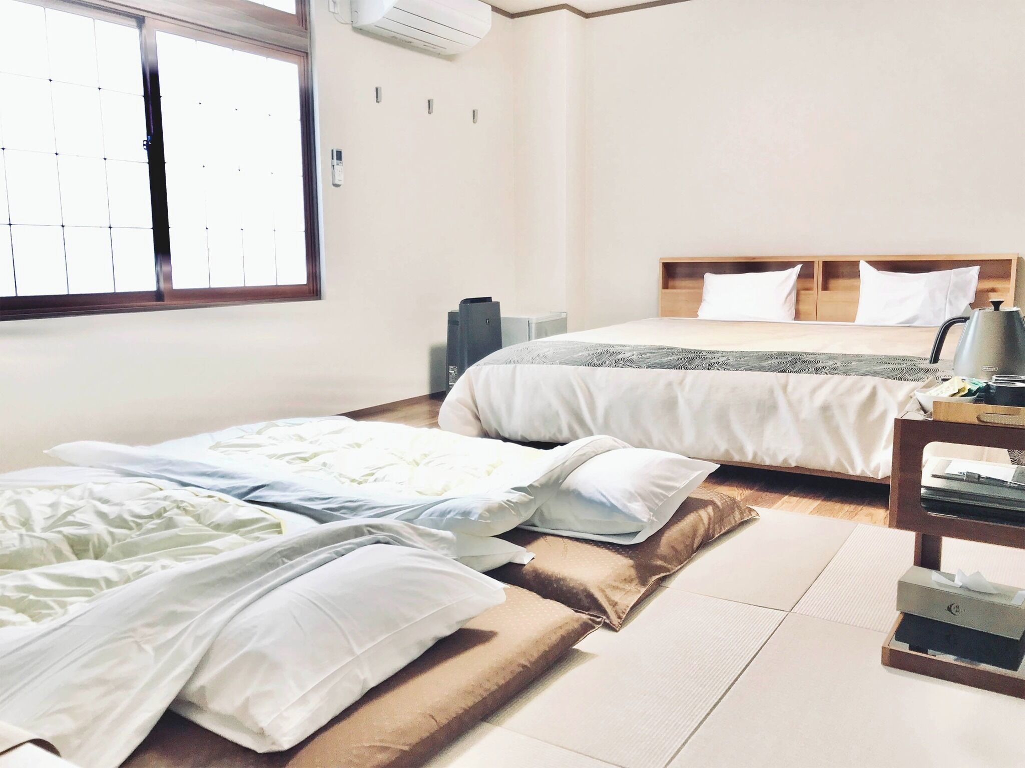 A hotel room with two beds, a wooden floor, a window, and a bedside table with a lamp.