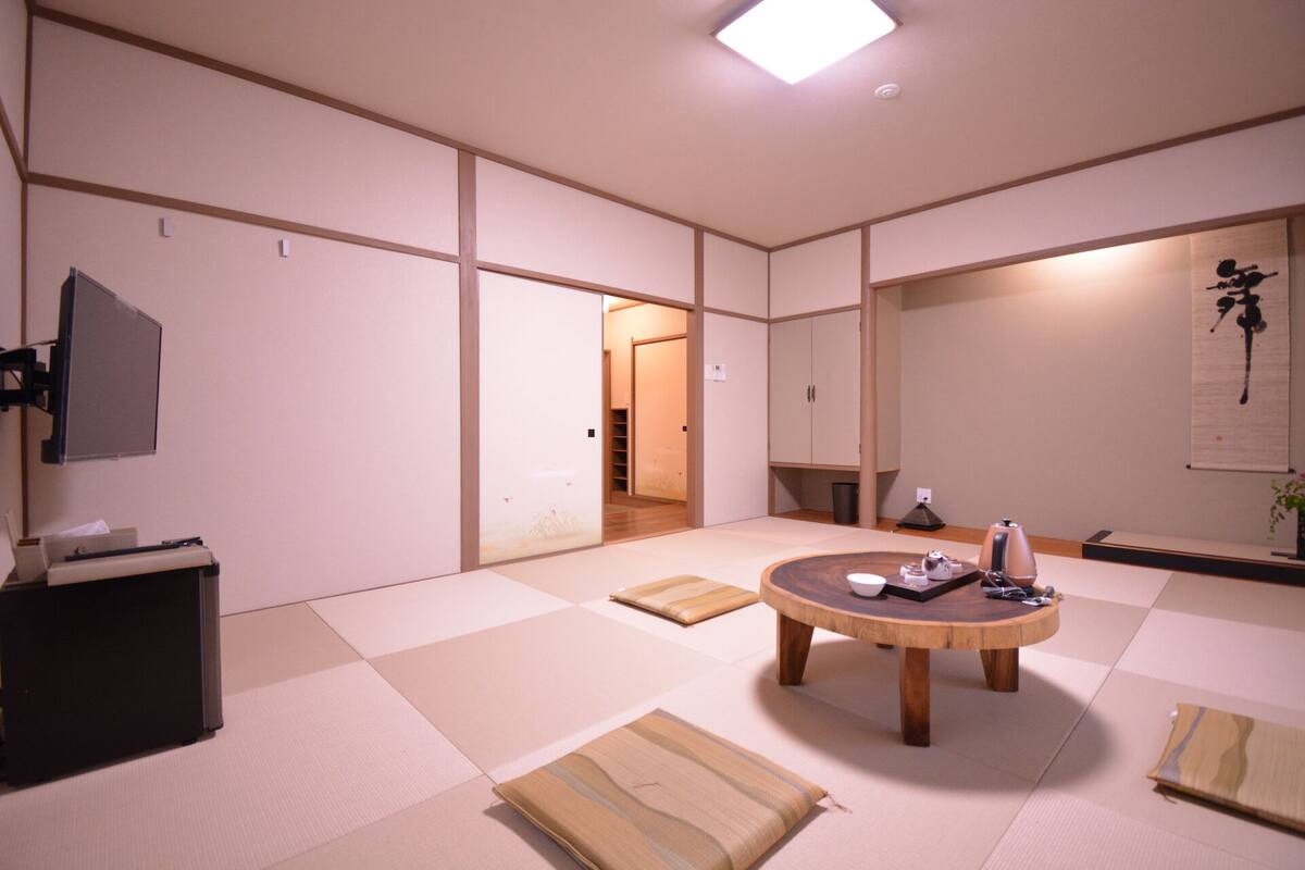 Een traditionele Japanse kamer met tatami vloerbedekking, een ronde houten tafel, een televisie en een schuifdeur naar een andere kamer.