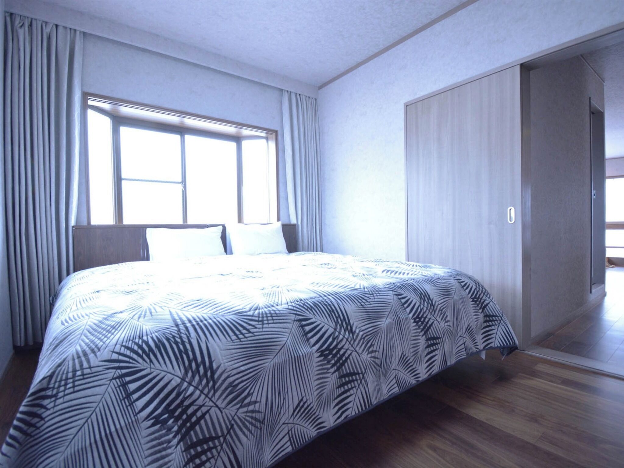 A hotel room with a large bed, a window with curtains, a wardrobe, and a door leading to another room.