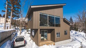 Una casa moderna de dos pisos con ventanas amplias, un automóvil estacionado adelante y nieve en el suelo.
