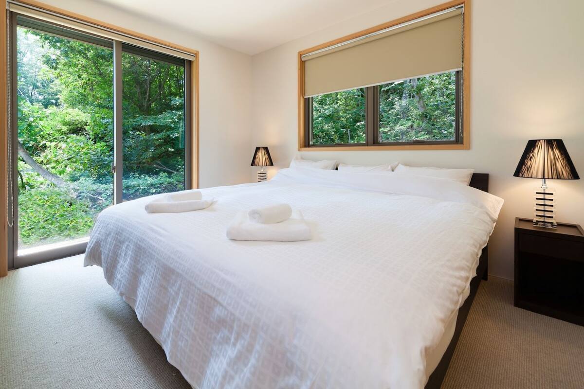 Camera d'albergo moderna con un letto grande, due lampade da comodino e vista sul verde attraverso ampie finestre a tutta parete.