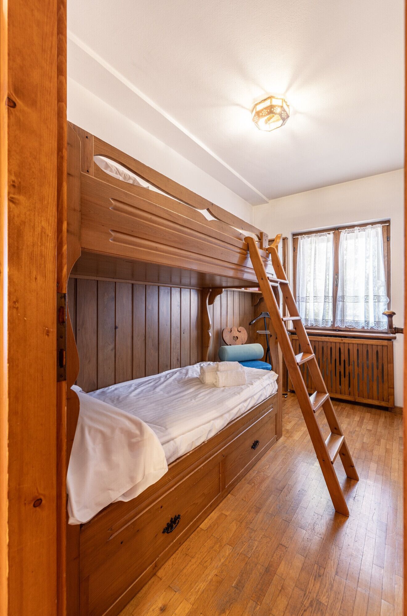 Una cama litera con escalera, suelo de madera y ventana con cortinas.