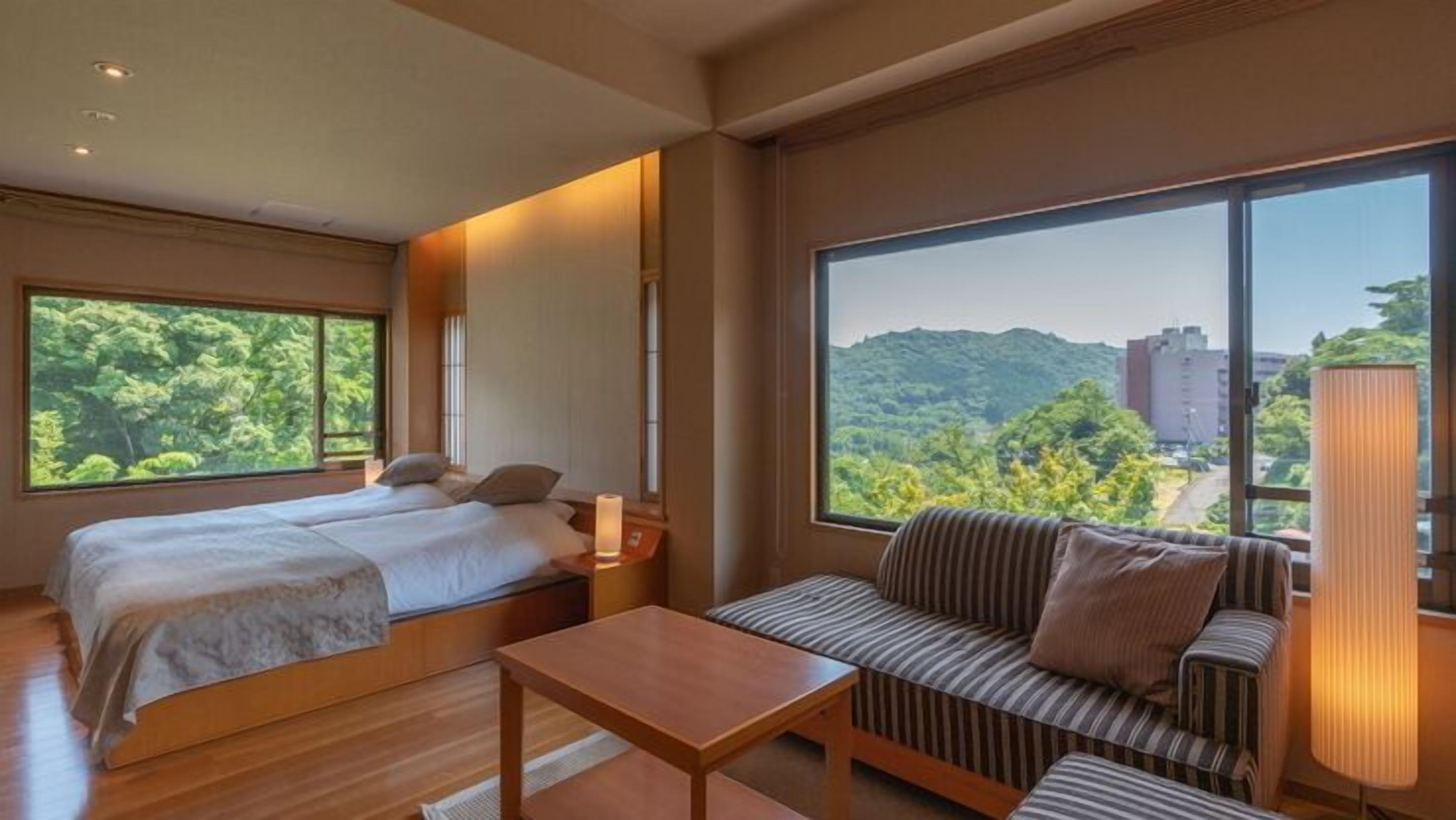 A hotel room with a bed, a sofa, a table, and large windows offering a view of greenery and buildings.