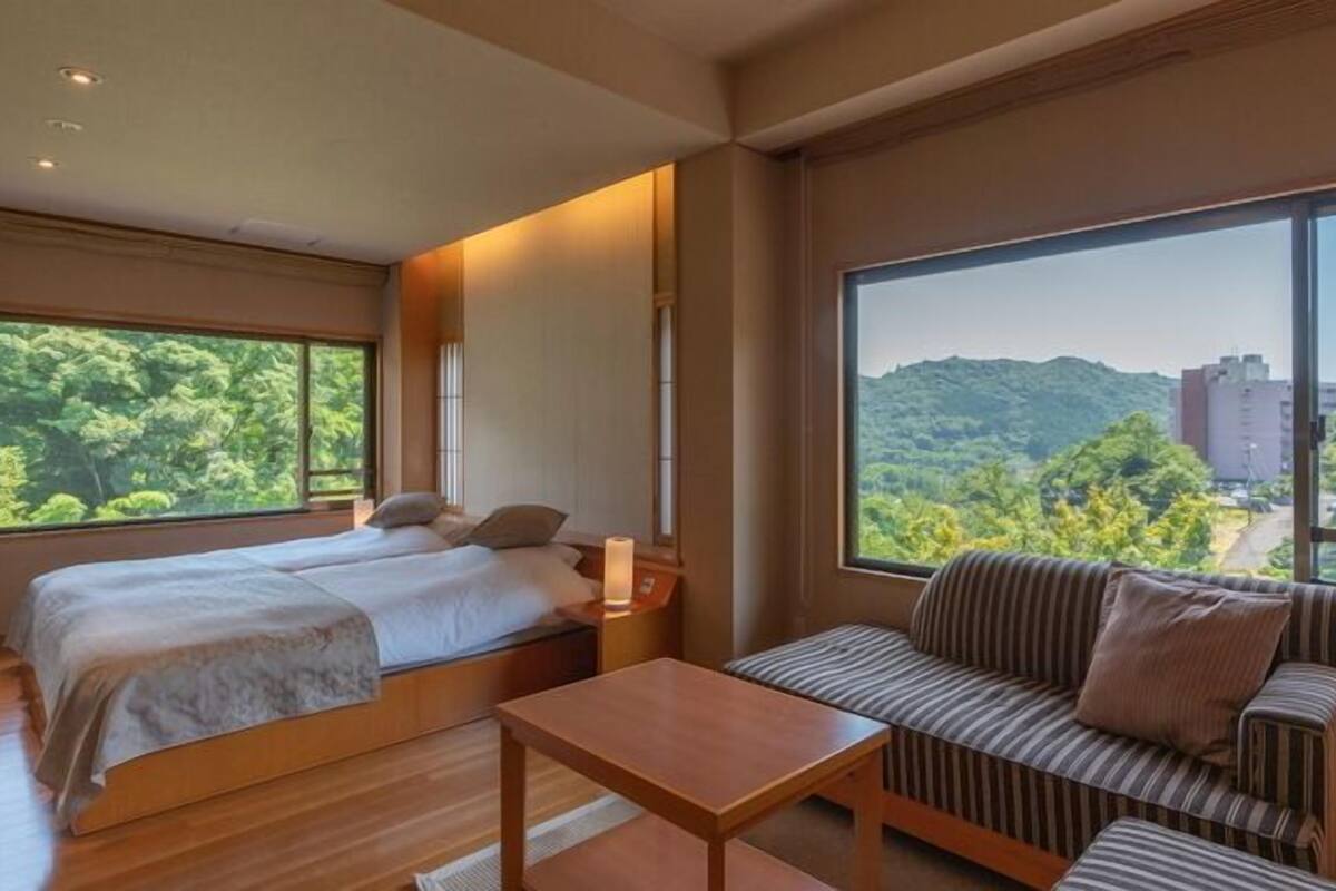 Ein Hotelzimmer mit Bett, einer Couch, einem Tisch und großen Fenstern mit Blick auf Grünflächen und Gebäude.