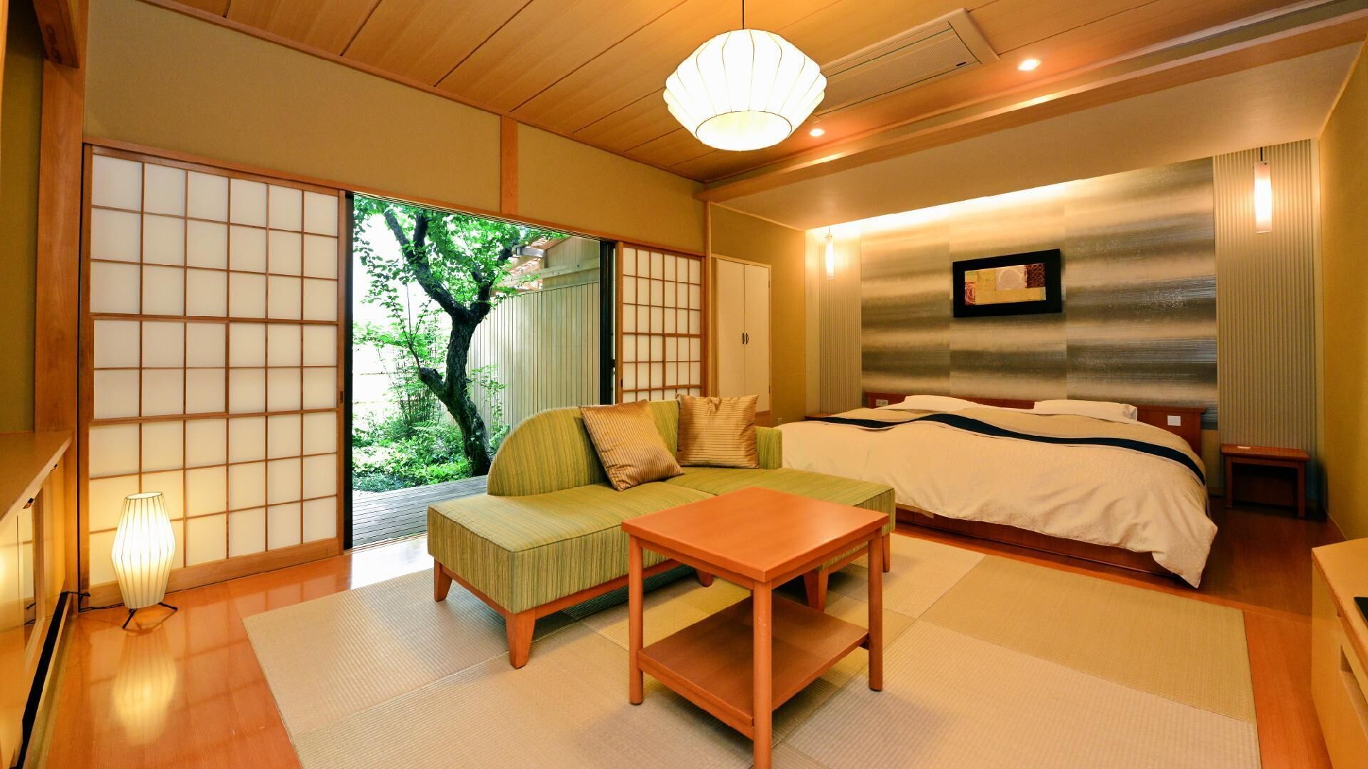 A traditional Japanese-style room with tatami flooring, a low wooden table, a sofa, and a bed with a large window offering a view of greenery.