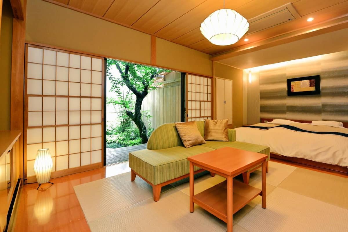 Ein traditioneller japanischer Raum mit Tatami-Boden, einem niedrigen Holztisch, einer Couch und einem Bett mit einem großen Fenster mit Blick auf Grünflächen.