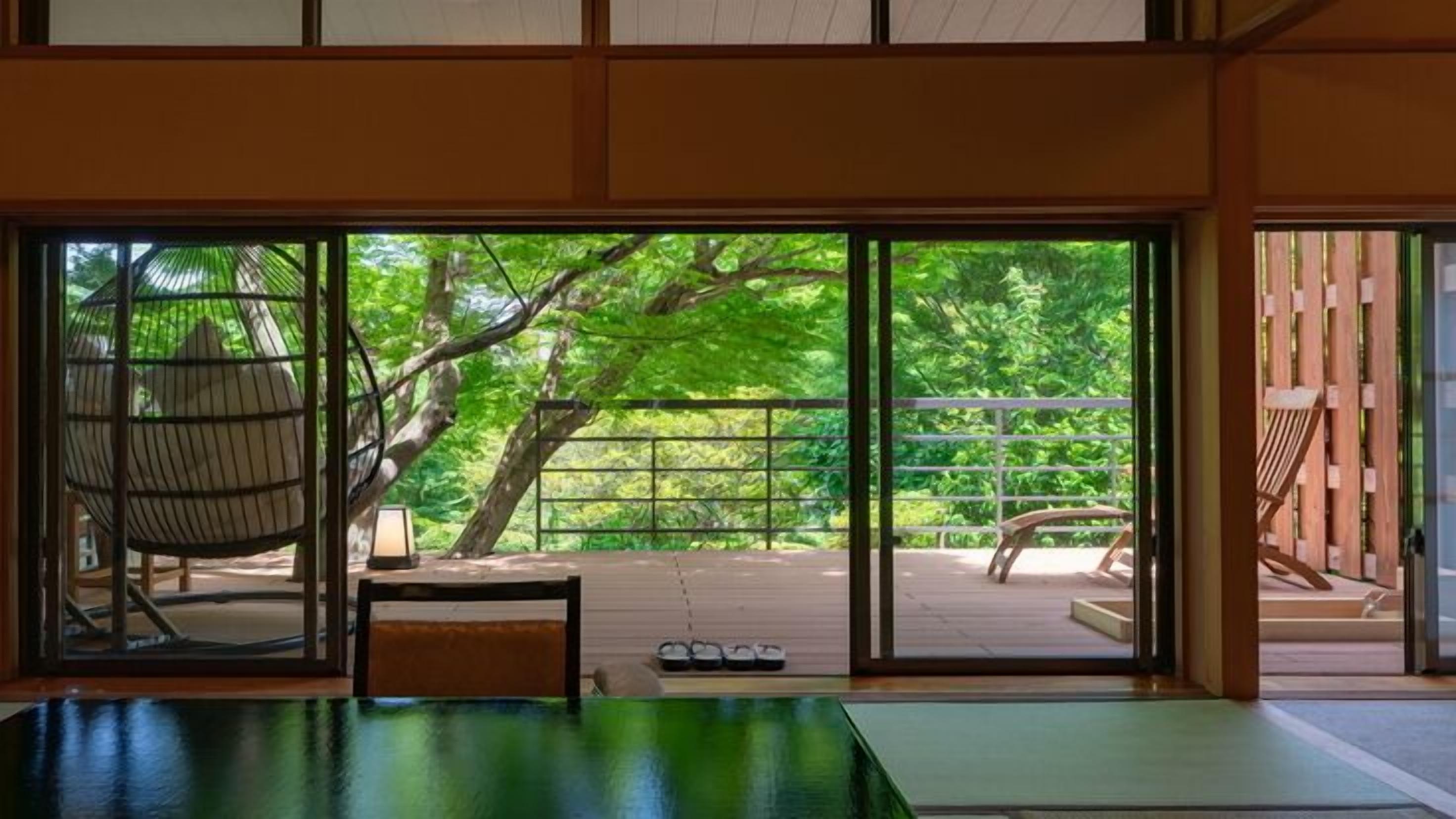 A room with a tatami mat floor, a low table, a chair, and a view of a wooden deck with a railing and a tree.