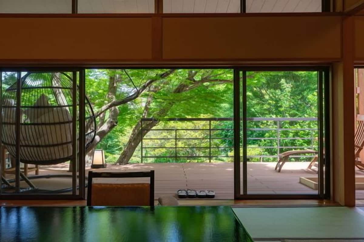 Ein Zimmer mit Tatami-Boden, einem niedrigen Tisch, einem Stuhl und Blick auf eine Holzterrasse mit Geländer und einem Baum.