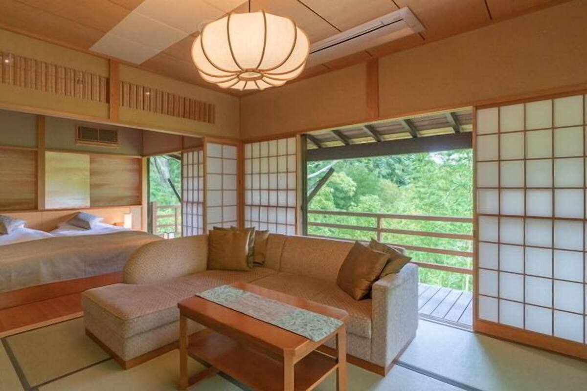 Ein traditionell japanisch gestalteter Raum mit Tatami-Böden, einem niedrigen Tisch, einer Couch und einem Bett mit Blick auf eine grüne Landschaft.