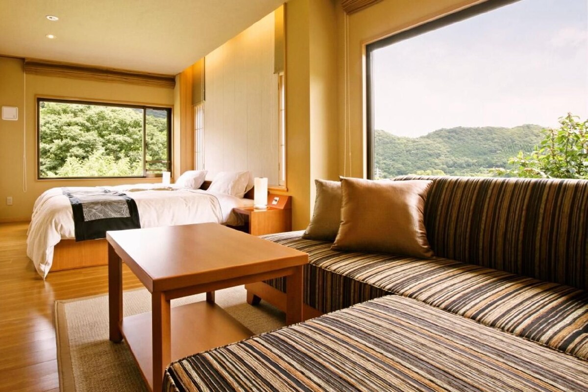 Een hotelkamer met een bed, een bank, een tafel en een groot raam met uitzicht op groen.