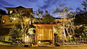 Un bâtiment de style japonais traditionnel, doté d’une porte en bois et entouré de bambous et d’autres plantes vertes.