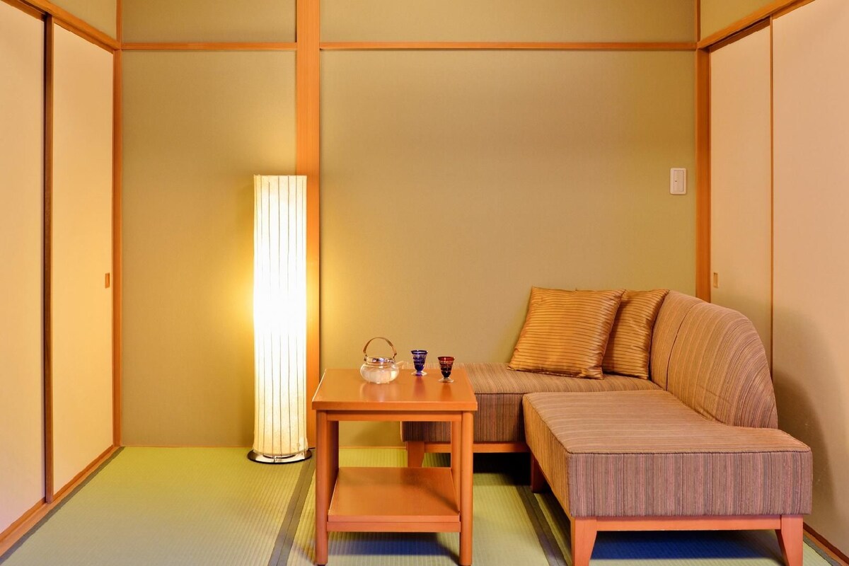 Een traditionele Japanse kamer met tatami vloeren, een lage houten tafel met een theeset, een bank met kussens en een hoge staande lamp.