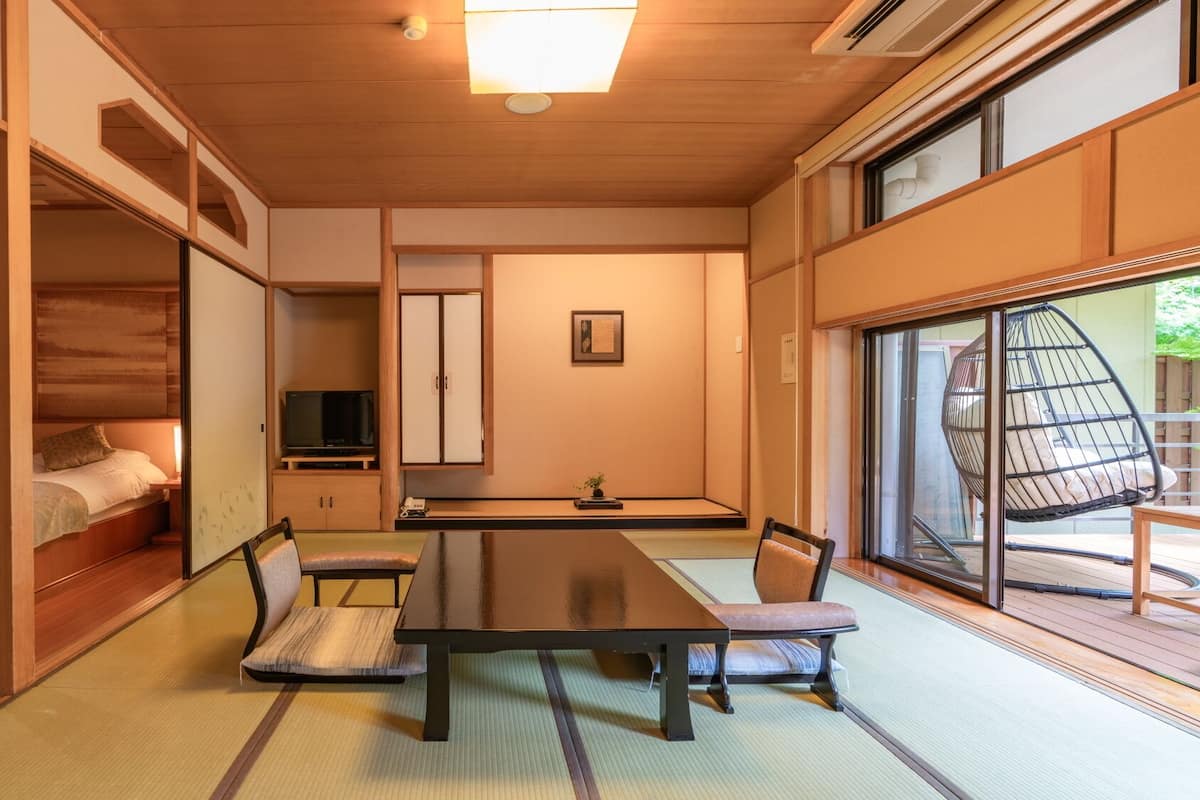 Ein traditioneller japanischer Raum mit Tatami-Bodenbelag, einem niedrigen Tisch mit Kissen, einem Fernseher, Schiebetüren und Blick ins Freie.