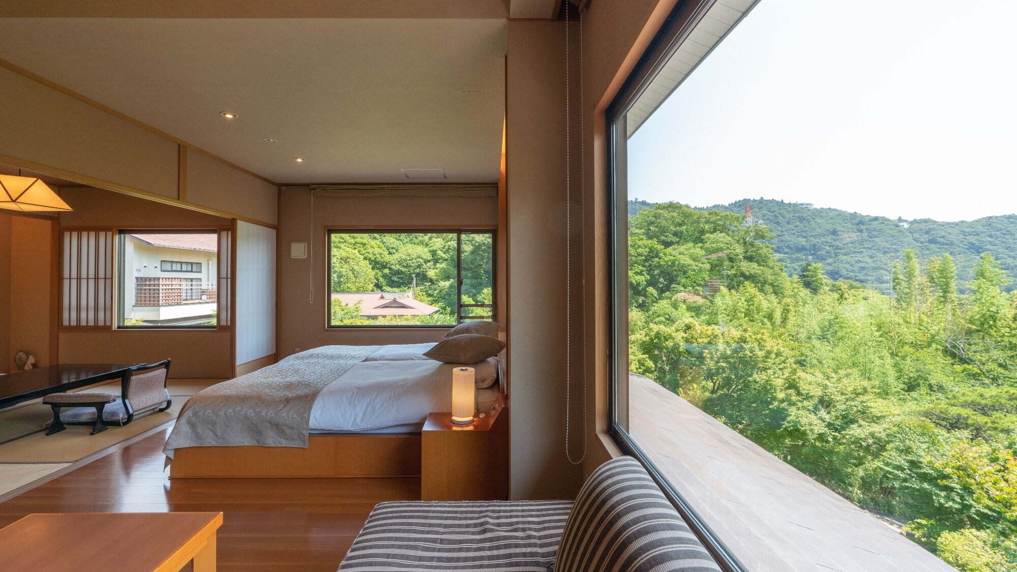 A bedroom with a large window, a bed, a bedside table with a lamp, a wooden desk, and a view of a forested hillside.