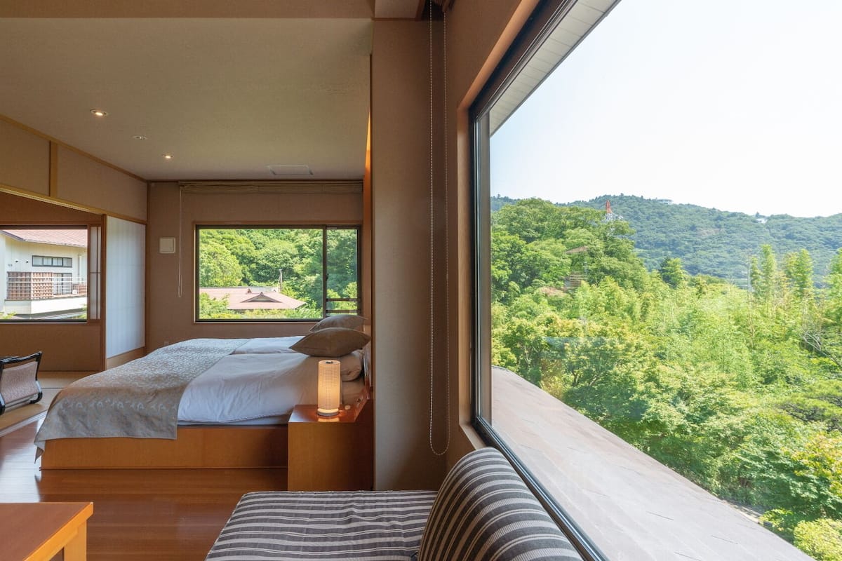 Ein Schlafzimmer mit großem Fenster, einem Bett, einem Nachttisch mit Lampe, einem Holztisch und Blick auf einen bewaldeten Hügel.