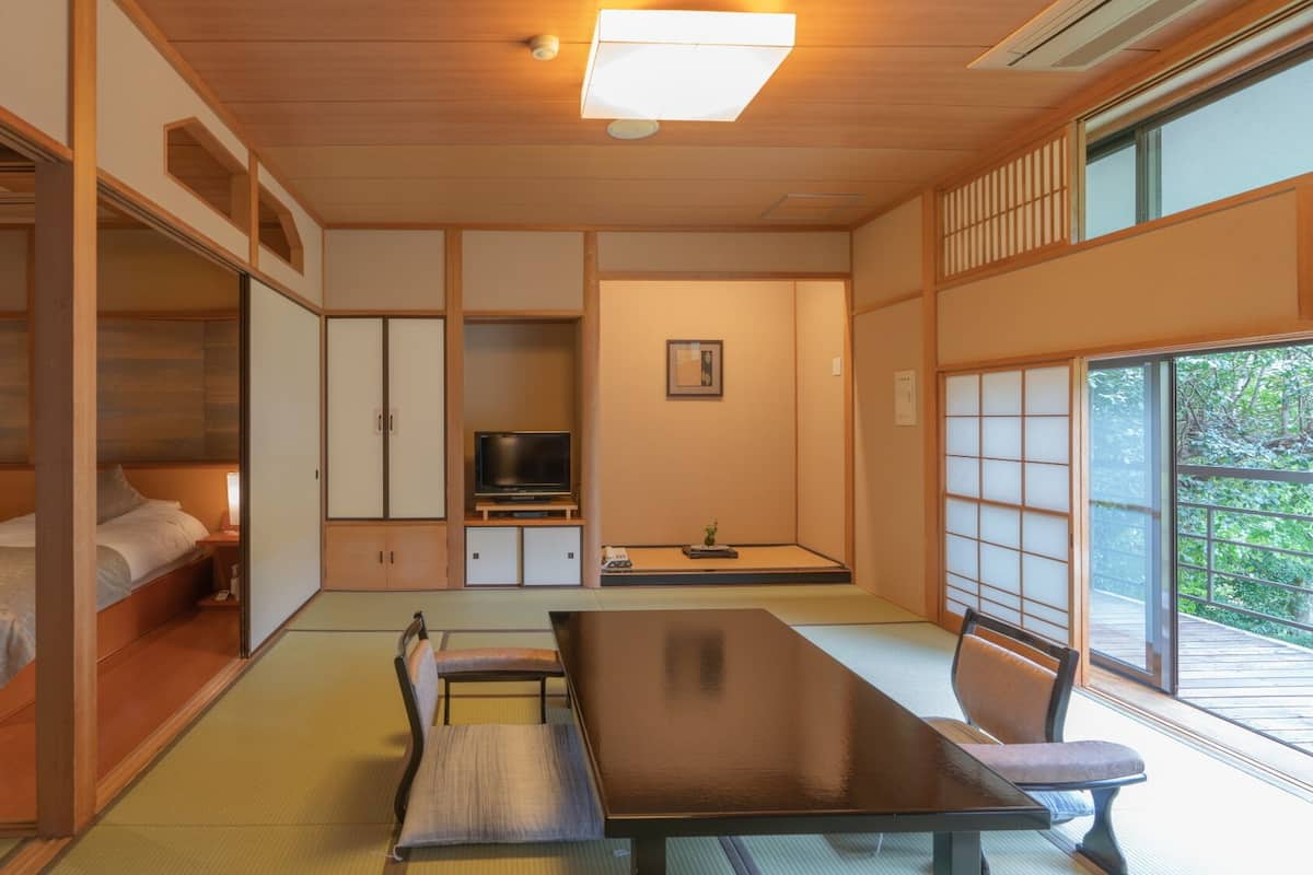 Ein traditioneller japanischer Raum mit Tatami-Boden, einem niedrigen Tisch mit Stühlen, einem Bett mit Kopfteil und Schiebetüren.