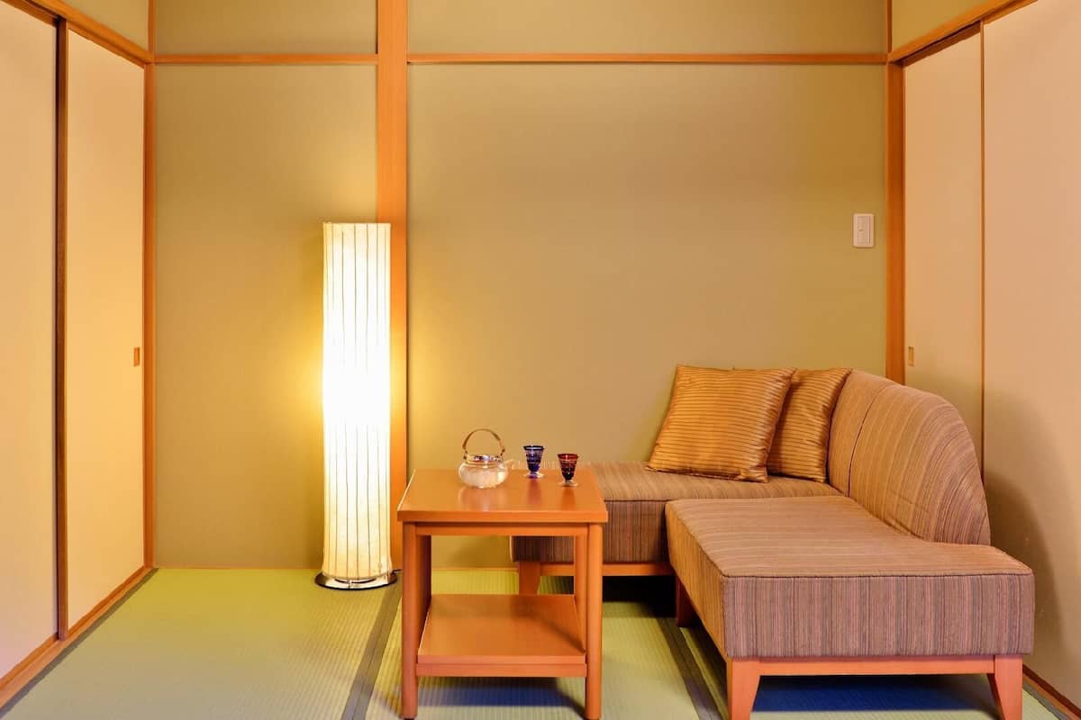 Ein traditioneller japanischer Raum mit Tatami-Boden, einem niedrigen Holztisch mit Teeservice, einer Couch mit Kissen und einer hohen Stehlampe.
