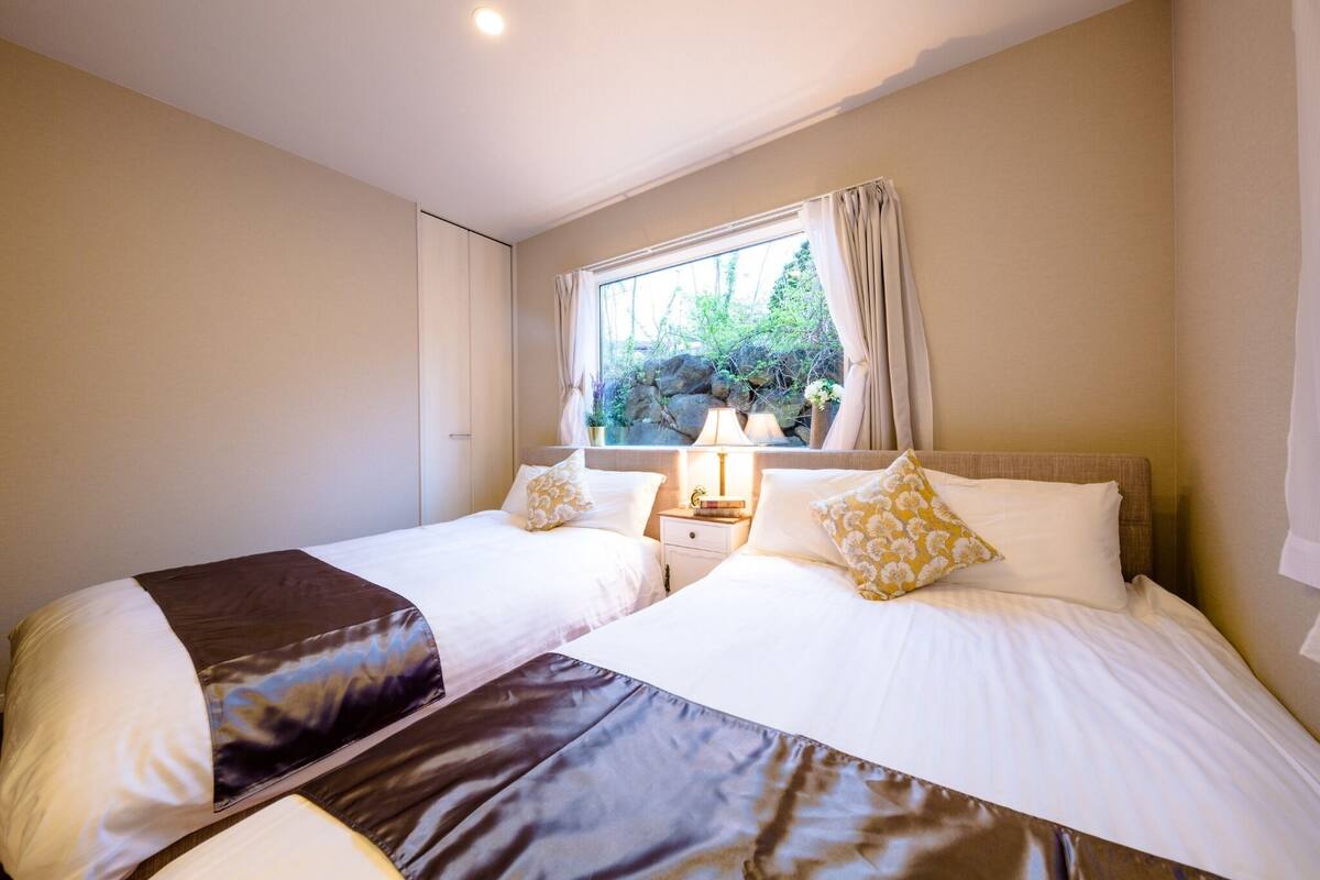 A hotel room with two beds, a window with curtains, a nightstand with a lamp, and a view of greenery outside.