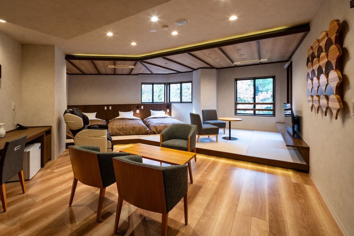 A modern hotel room with a wooden floor, a desk with a chair, a massage chair, a bed with two pillows, a small table, and a view of trees through large windows.