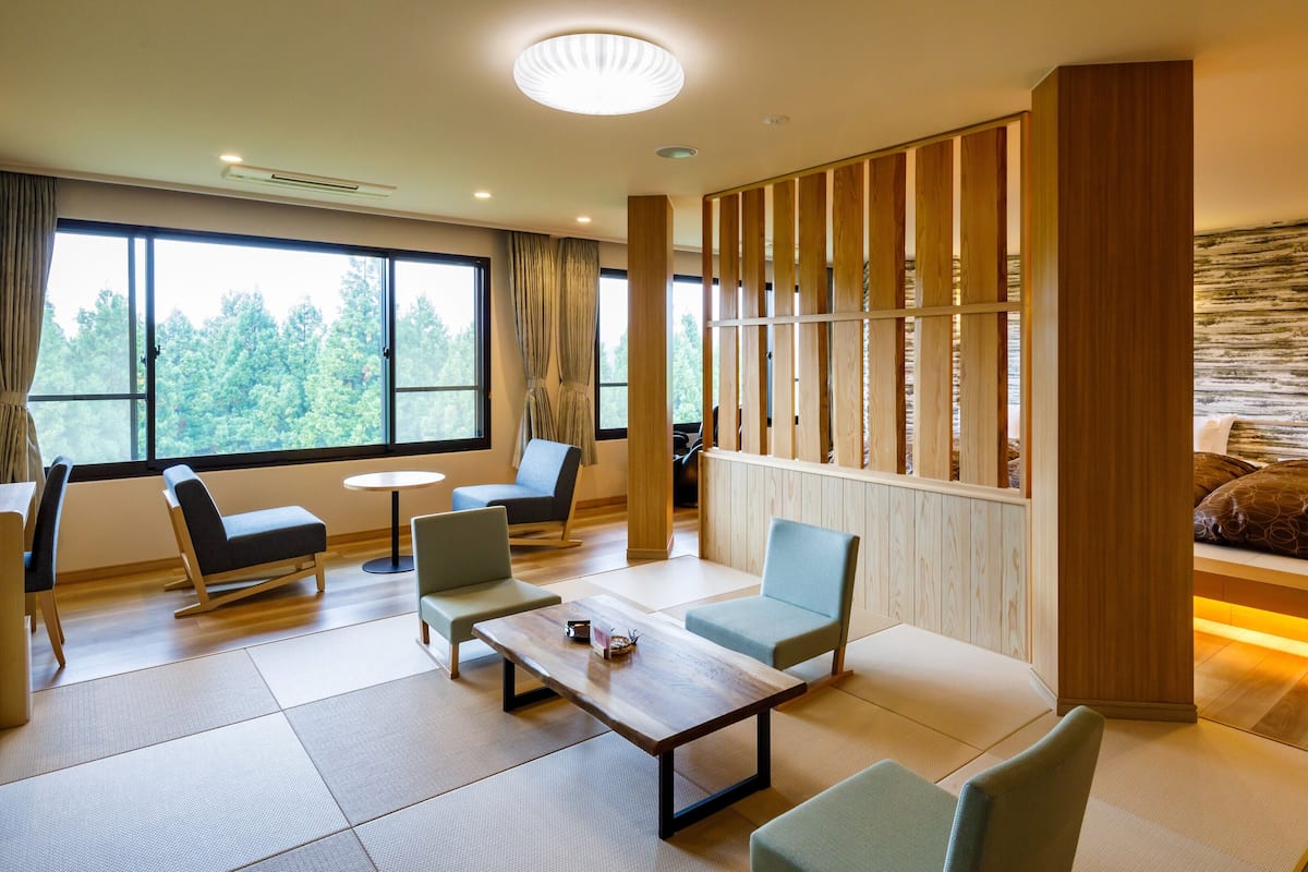 A modern hotel room with a large window, a bed with a wooden headboard, a seating area with chairs and a table, and a decorative wooden partition.