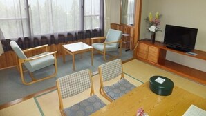 Ein Zimmer mit Tatami-Matten, einem Fernseher, einem Schrank und einem Fenster mit Vorhängen.