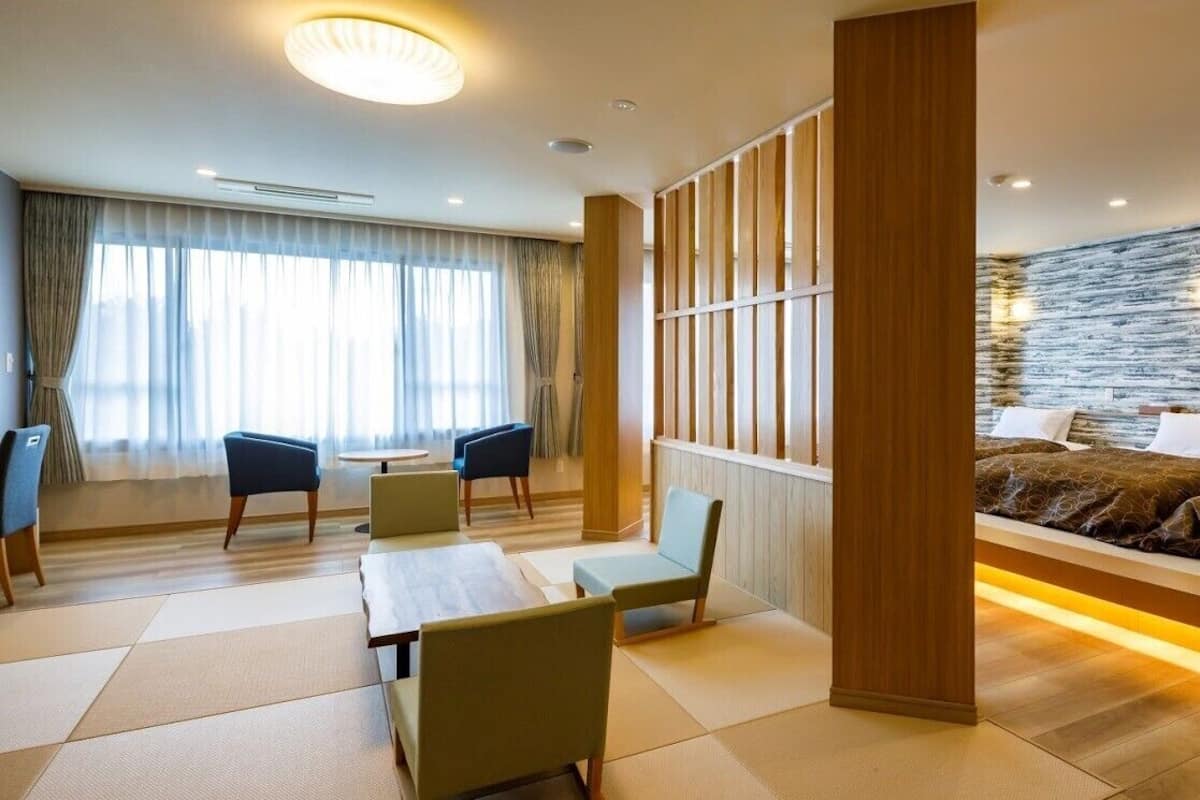 A hotel room with a sofa bed, a dining area with chairs and a table, and a large window with curtains.