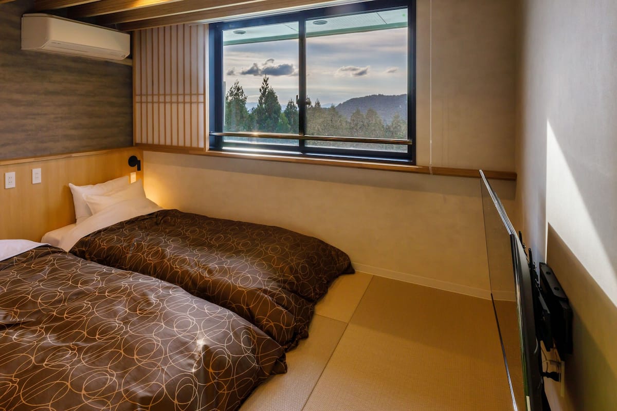 A hotel room with two beds, a window with a view of trees and hills, a wall-mounted air conditioner, and a wooden headboard.