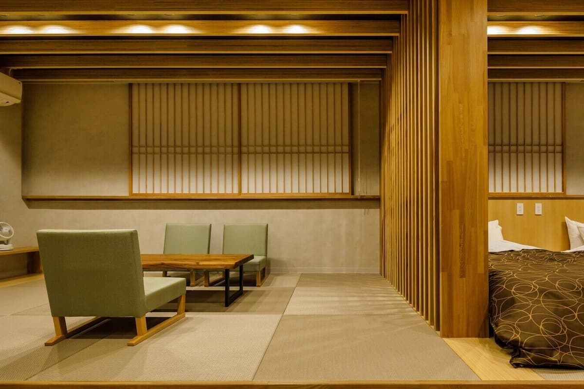 A room with tatami flooring, a low wooden table, green chairs, a bed with a patterned bedspread, and sliding shoji screens.