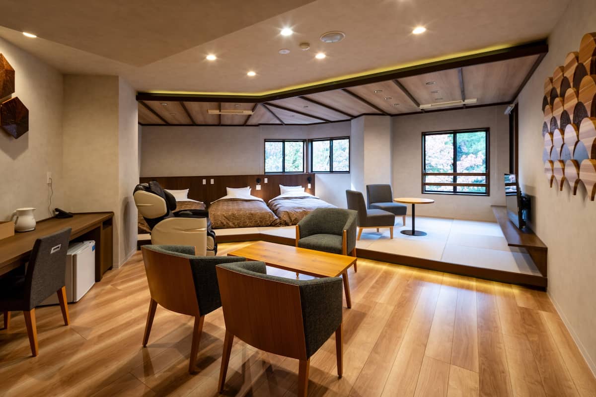 A modern hotel room with a wooden floor, a desk with a chair, a massage chair, a bed with two pillows, a small table, and a window with a view of trees.