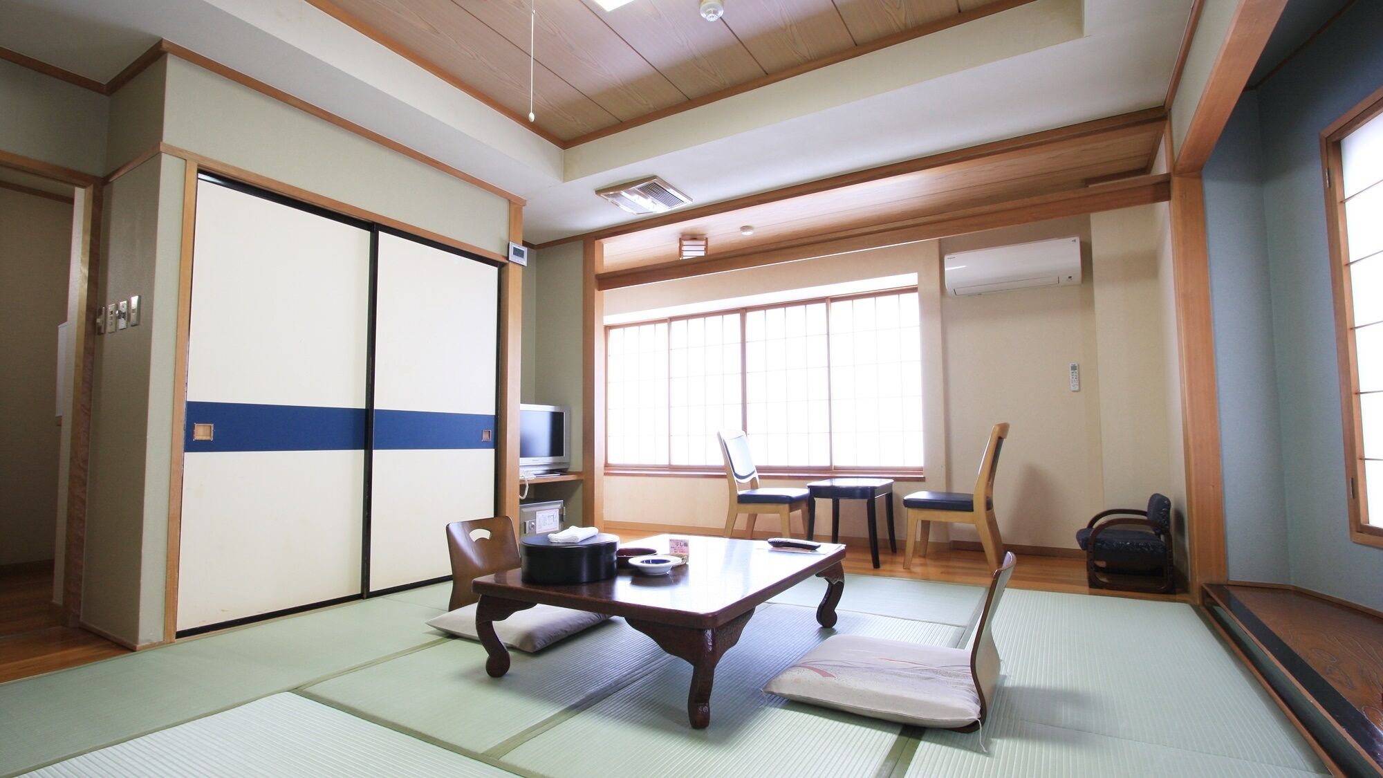 Ein traditioneller japanischer Raum mit Tatami-Bodenbelag, einem niedrigen Tisch, Stühlen, einem Fernseher und Schiebetüren.