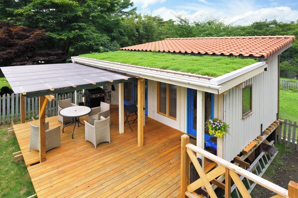 Un edificio pequeño de un solo piso con techo plano, una terraza con muebles de exterior y un patio cubierto de césped.