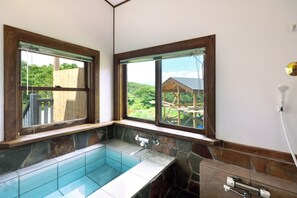 A bathroom with a built-in bathtub, stone walls, and large windows offering a view of a wooden deck and greenery. - Kurumi Terrace (Tsumagoi)