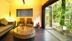 Un interior moderno con una mesa de centro redonda de vidrio, un sofá y una silla. Hay una ventana con persianas y vistas a zonas verdes al exterior.